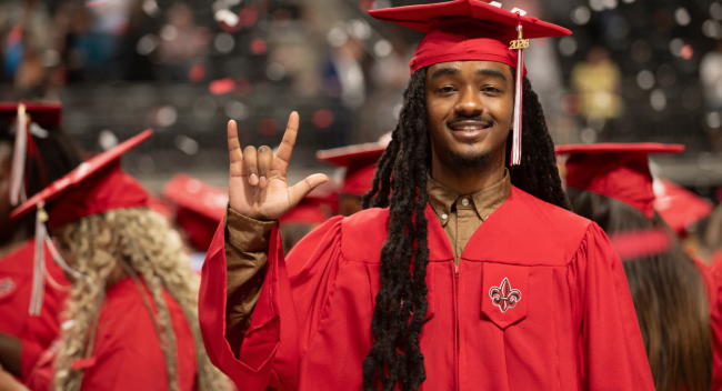 Graduate of the online health services administration degree program smiles in his cap and gown, with confetti falling in the background.